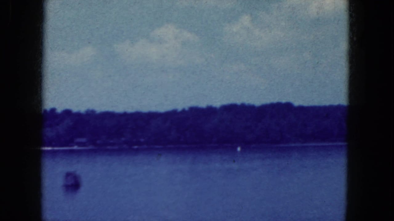 1964 8mm home-movie frame from the Niagara Falls, New York metadata showing calm lake water, distant trees, pale sky, and a small buoy.
