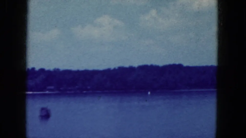1964 8mm home-movie frame from the Niagara Falls, New York metadata showing calm lake water, distant trees, pale sky, and a small buoy.