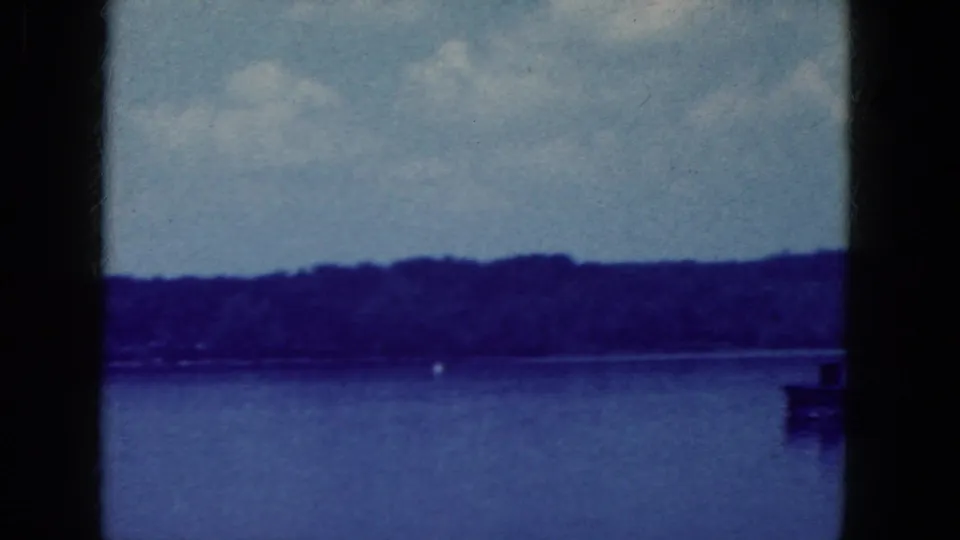 Blue-cast 1964 8mm frame showing a calm lake, wooded far shore, small buoy, and dark vertical frame divisions.