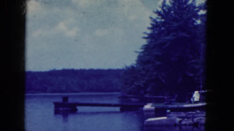 Blue-toned 1964 8mm frame showing a lakeside dock, dense trees, calm water, and distant shore in the Niagara Falls, New York metadata.
