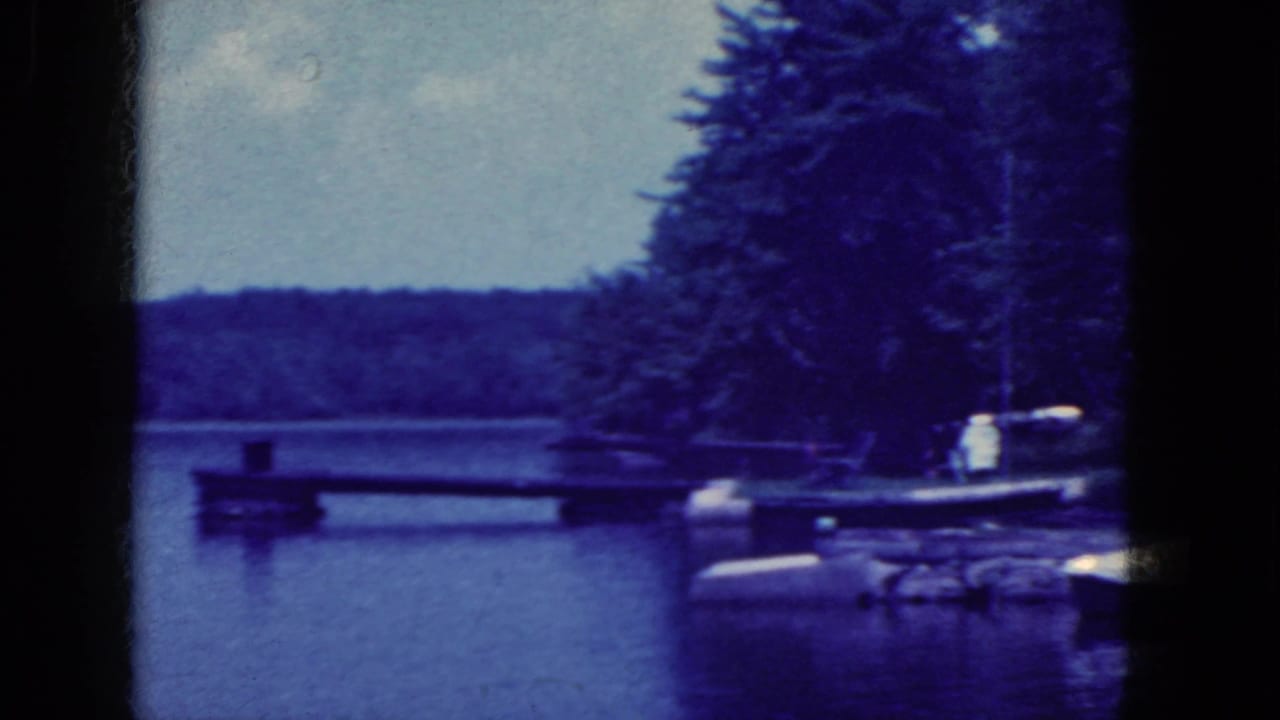 1964 8mm frame from Niagara Falls, New York, showing a calm lake, dock, trees, and distant shoreline.