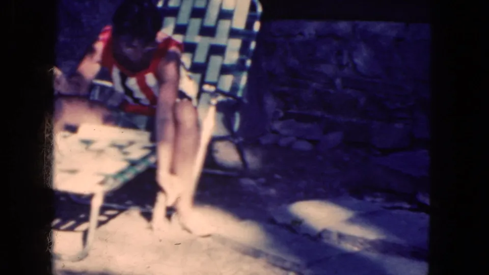 1964 8mm frame of a woman in a striped swimsuit sitting by a stone wall as a small squirrel stands near her feet.