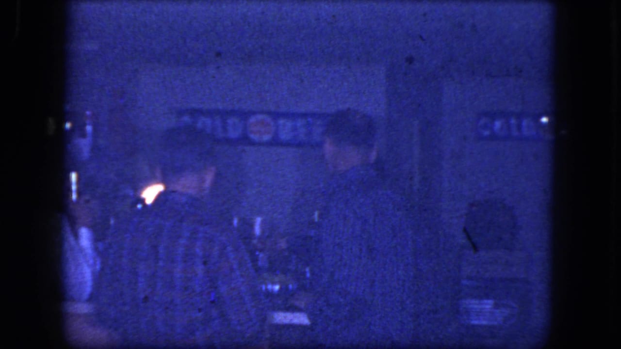 1964 Niagara Falls home-movie frame showing men seated at a dim bar counter, with a COLD BEER sign and drinking glasses behind them.
