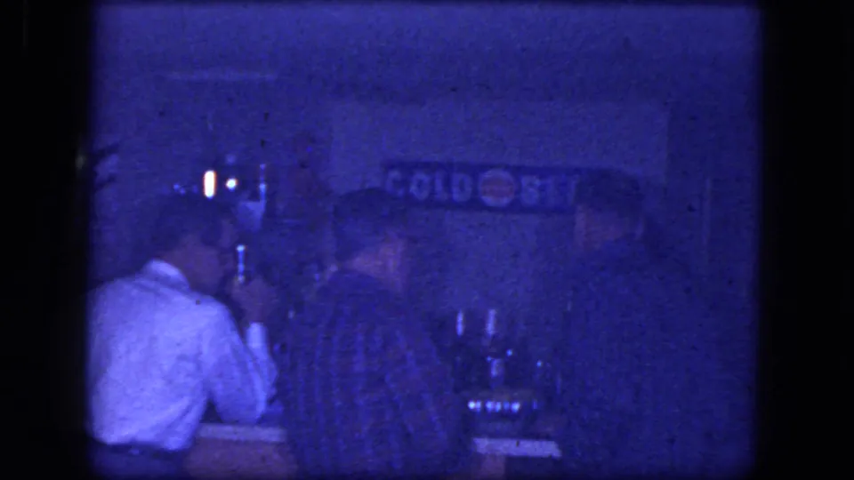 1964 Niagara Falls bar interior with men at the counter, one in a white shirt and another in plaid, under dim blue-tinted light.
