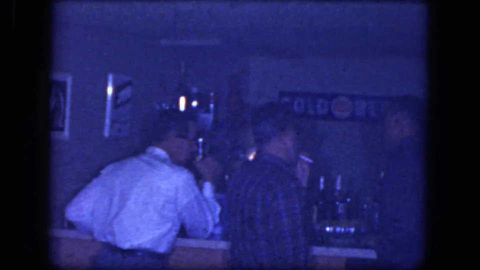 Blue-tinted 8mm frame from a Niagara Falls bar showing a man leaning on the counter while another appears to smoke beside him.