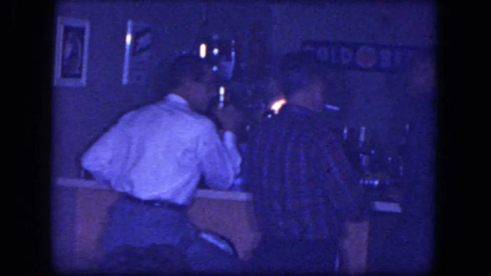 1964 Niagara Falls home-movie frame of men seated at a dim bar counter, with a lit cigarette, wall sign, and glasses in view.