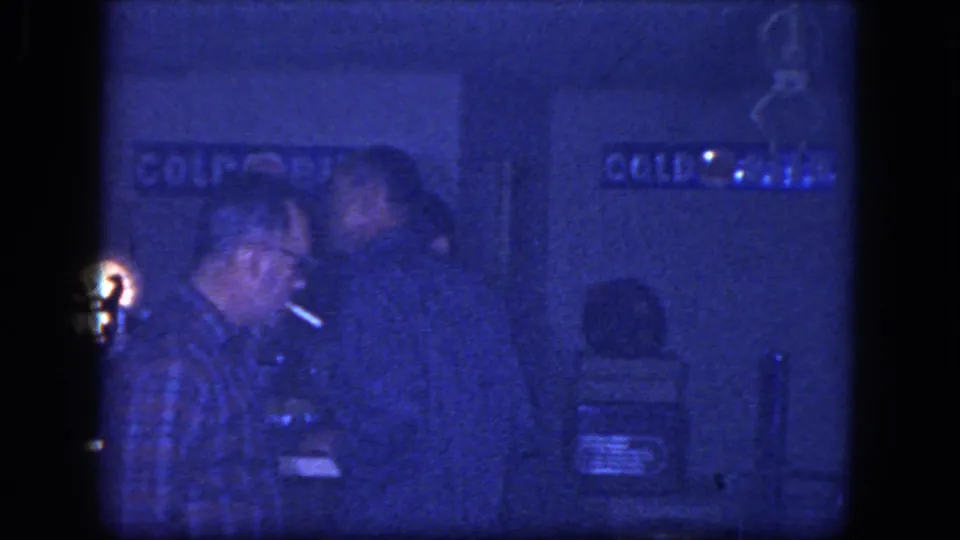 A cigarette flame briefly illuminates an adult man's face in the dark bar interior from the 1964 Niagara Falls home movie.