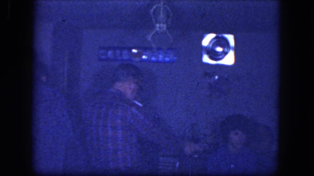 1964 Niagara Falls home-movie frame showing adults in a dim private room, with a standing man in plaid near seated party guests.