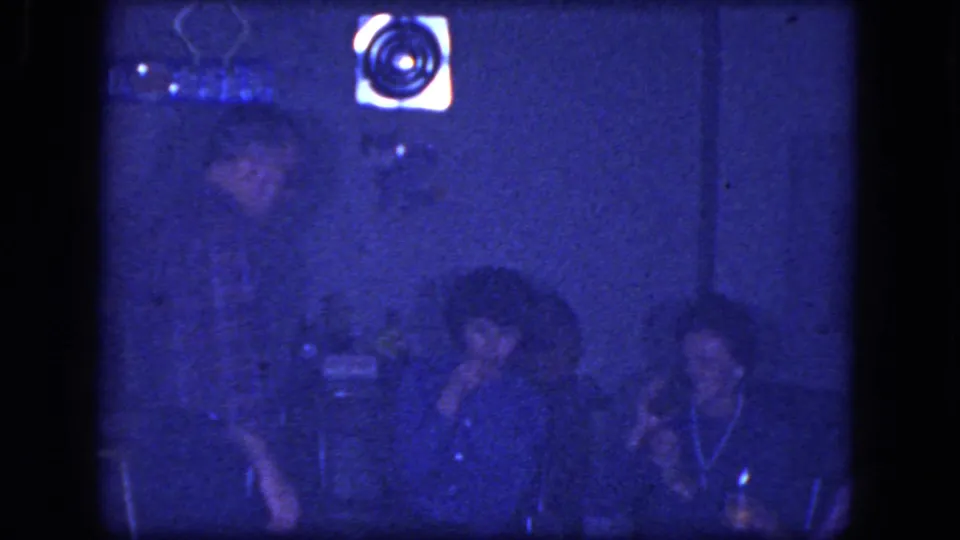 1964 Niagara Falls home-movie frame of seated adults socializing indoors, including visible smoking in dim blue light.
