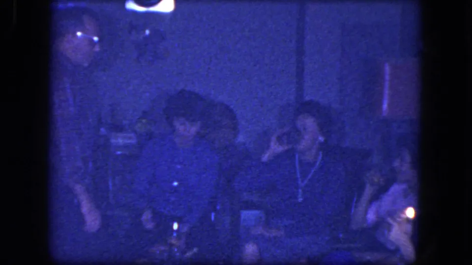 1964 Niagara Falls home-movie frame showing adults at an indoor party with cigarettes, drinks, and low household lighting.