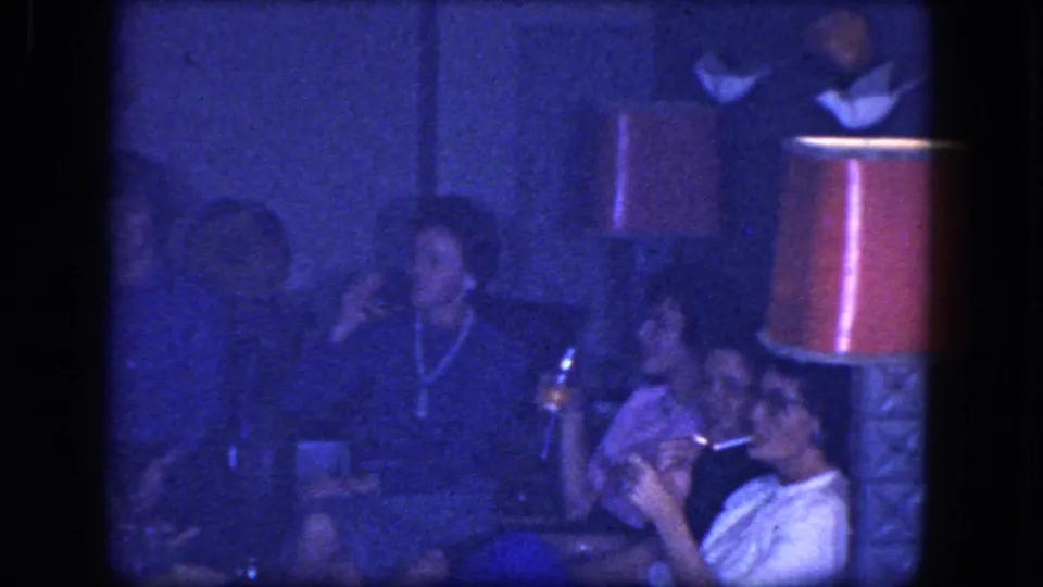 1964 Niagara Falls home-movie frame of adults seated in a private room beneath red lampshades, with smoking and bottles visible.