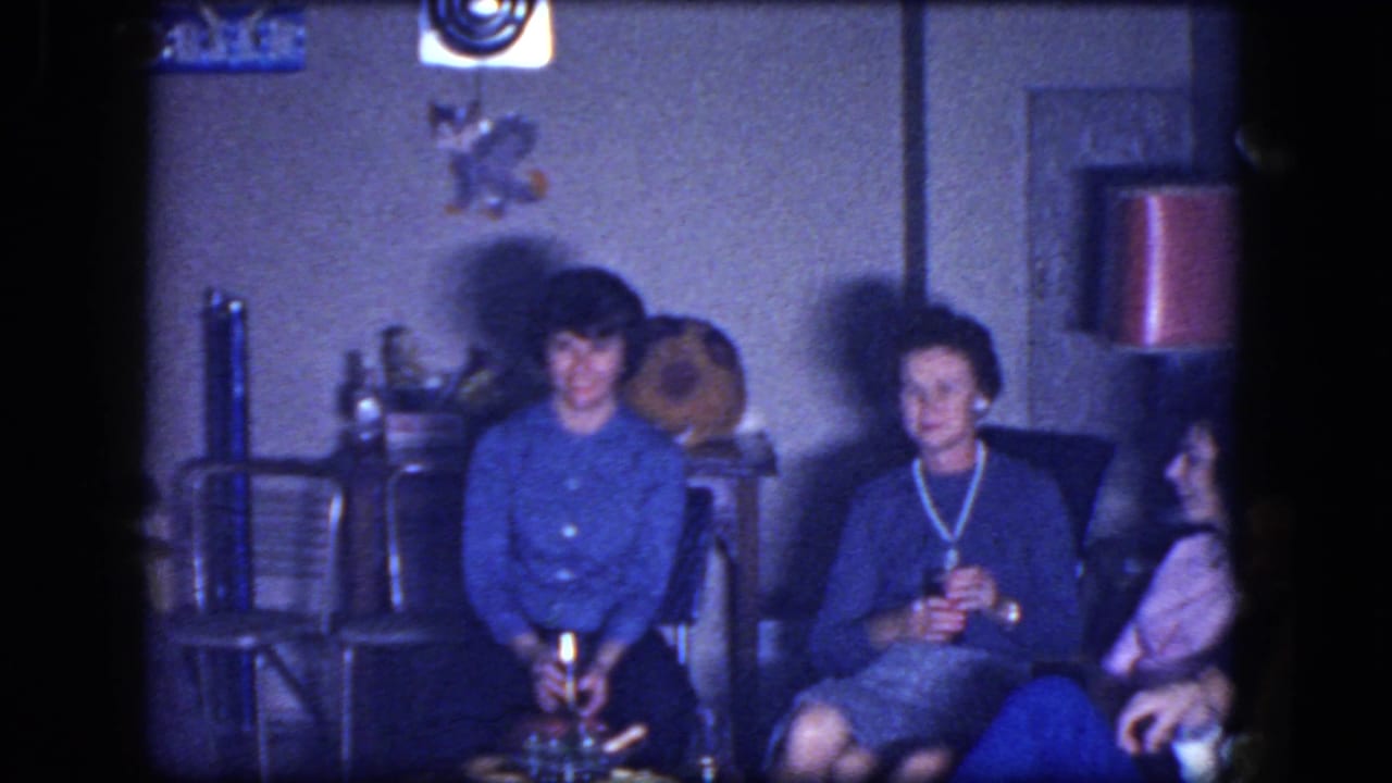 1964 home-movie frame of women seated in a private living room, with a small table, wall decor, and stacked chairs nearby.