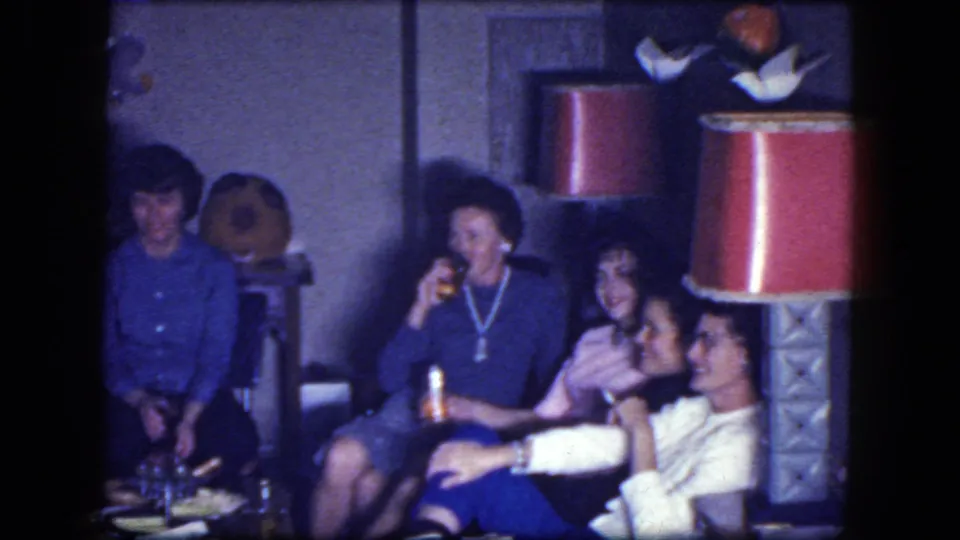 A home-movie frame shows women socializing in a private room, with a red-shaded lamp and small table in the foreground.