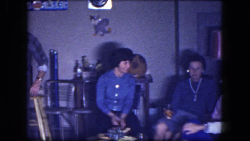 Women sit in a private living room in 1964 footage, with chairs, wall decor, and a red lampshade visible around them.