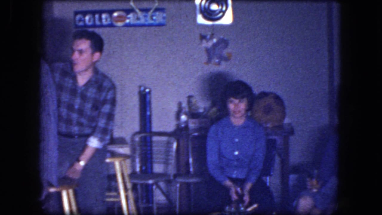 Men and a seated woman in a dim basement room with stools and beer signs on the wall.