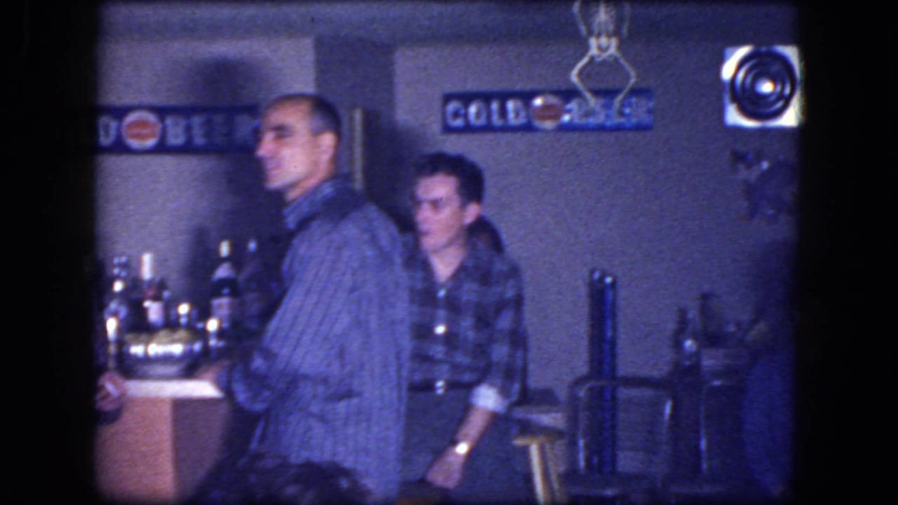 1964 Niagara Falls home-movie frame with men standing by a small basement bar, bottles, stools, and beer signs.
