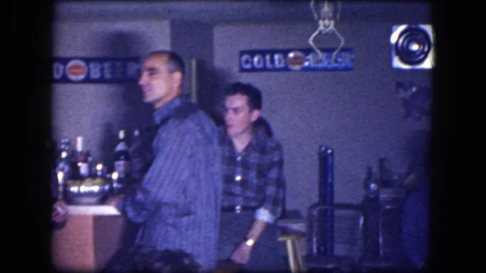 1964 Niagara Falls home-movie frame with men standing by a small basement bar, bottles, stools, and beer signs.