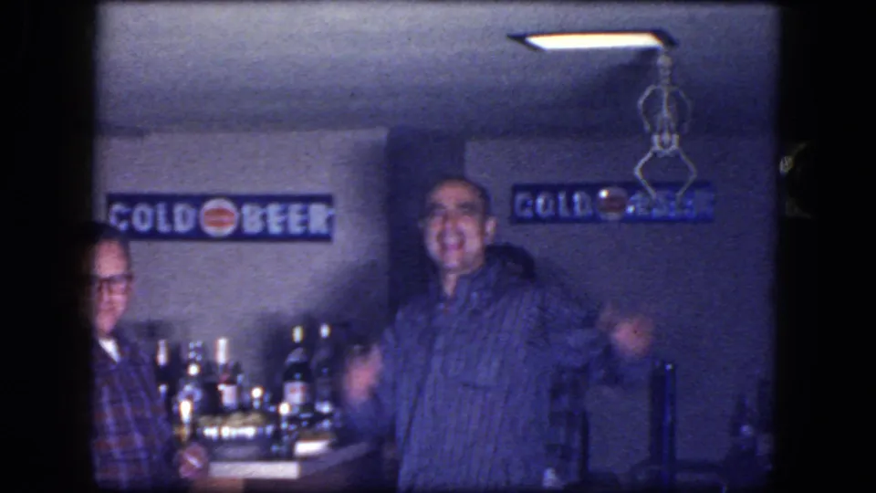 1964 Niagara Falls basement party frame showing men near a home bar with bottles, signs, and dim overhead lighting.