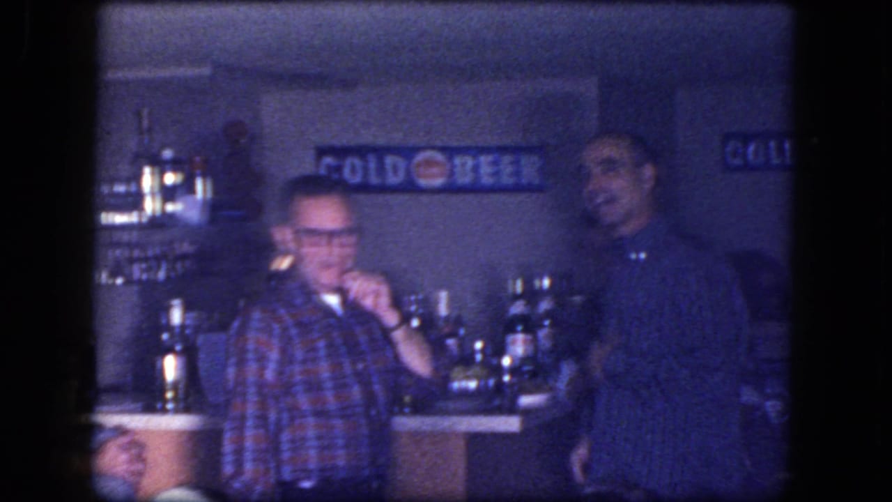Two adult men at a bar counter with liquor bottles and a COLD BEER sign behind them.