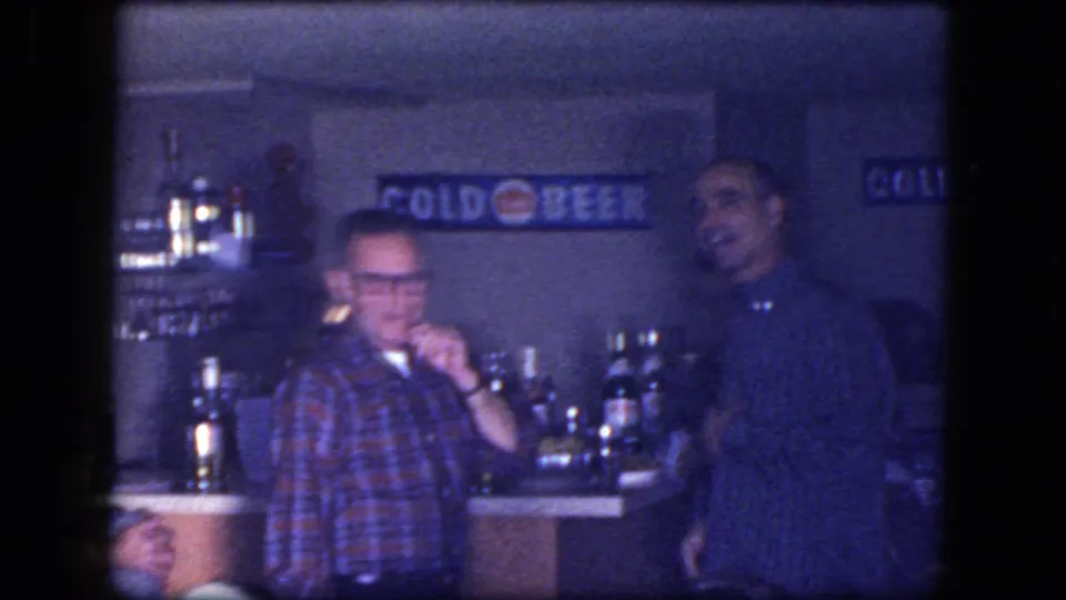 1964 Niagara Falls home-movie frame showing two adult men near a bar counter with bottles, chrome fixtures, and a COLD BEER sign.