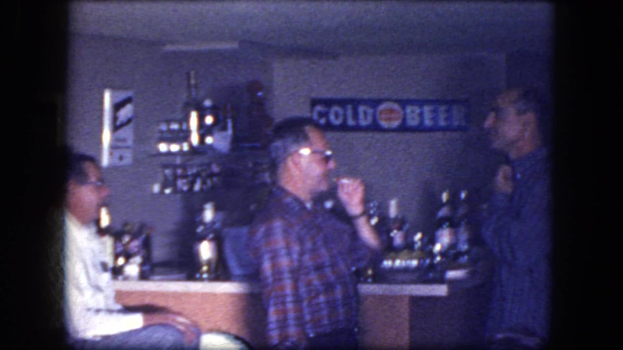 Three adult men at a bar, with one seated in a white shirt and two others facing each other.