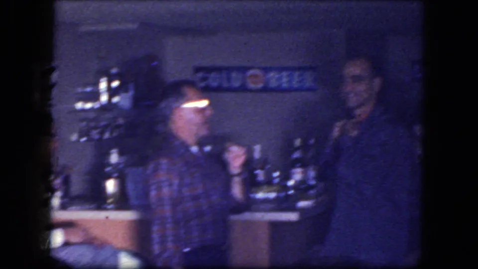 Blue-tinted 8mm frame from a 1964 Niagara Falls bar scene, with two men, counter bottles, and a COLD BEER sign in view.