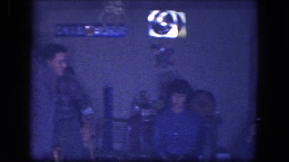Very dim 1964 Niagara Falls bar interior frame with seated figures, wall signage, bottles, and heavy blue 8mm exposure.