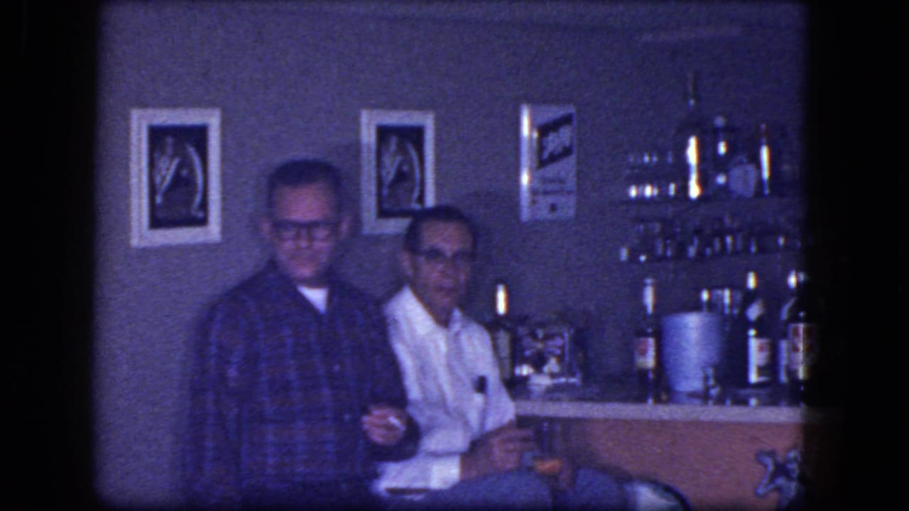1964 home-movie frame from Niagara Falls, New York, showing two adult men seated near bottles and glassware in a dim bar interior.