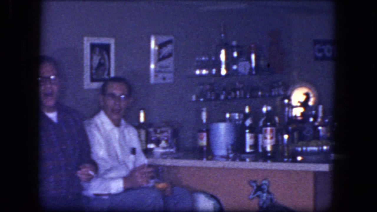 Two seated men face the camera near a bar shelf with bottles and a glowing lamp.