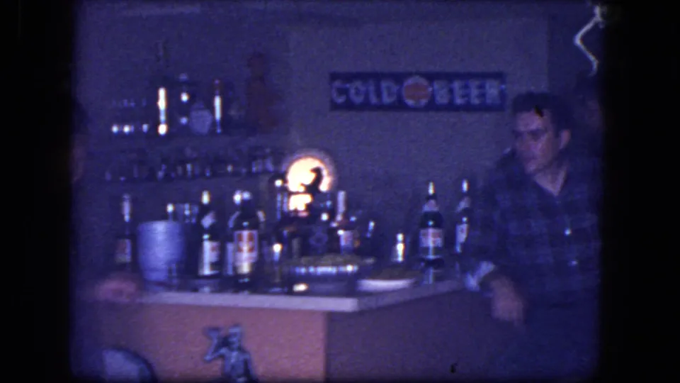 1964 Niagara Falls home-movie frame with an adult man seated by a bar counter, liquor bottles, glassware, lamp, and Cold Beer wall sign.
