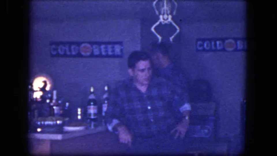 1964 home-movie frame from Niagara Falls showing an adult man seated near a stocked bar, dim lamp, bottles, and visible Cold Beer signs.