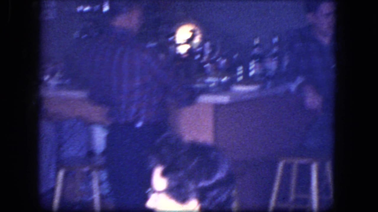 1964 Niagara Falls home-movie frame showing a woman in a white shirt and dark glasses seated in a blue-tinted bar interior.