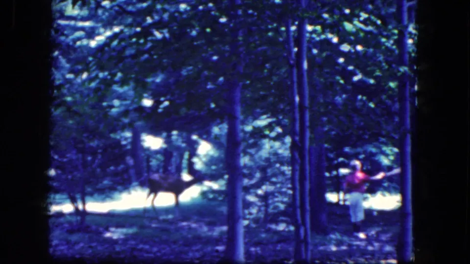 A person in red and light pants passes near the right edge as a deer remains visible in the wooded 1964 home-movie frame.