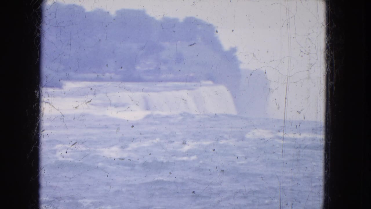 1964 8mm Niagara Falls view from above, showing heavy water flow, mist, distant cliff shapes, scratches, and dark gate edges.