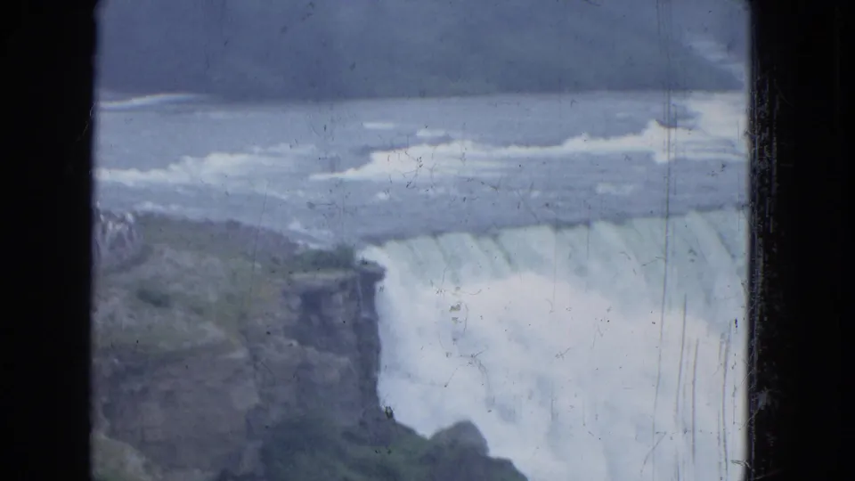 1964 8mm frame showing Niagara Falls from across the gorge, with rushing river water and mist near the drop.