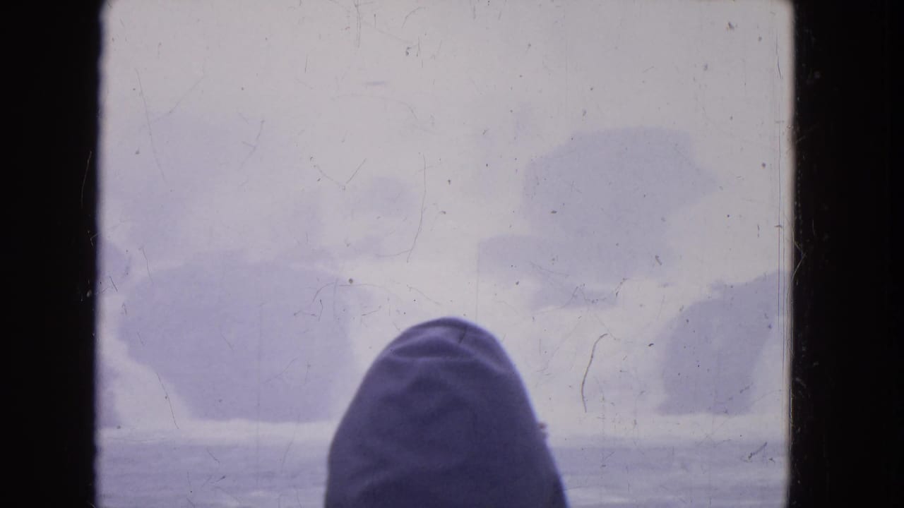 1964 8mm home-movie frame near Niagara Falls showing a hooded person viewed through a boat window against foggy winter water.