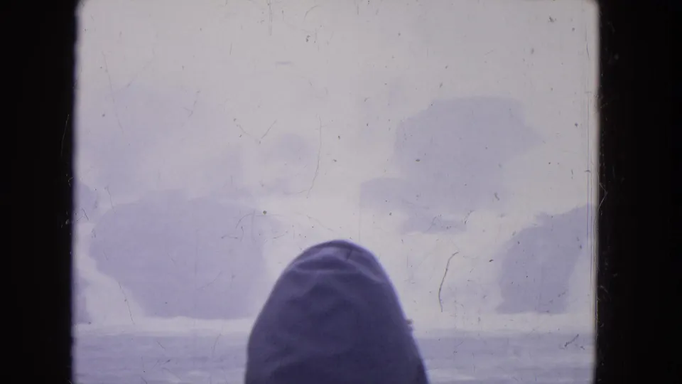 1964 8mm home-movie frame near Niagara Falls showing a hooded person viewed through a boat window against foggy winter water.
