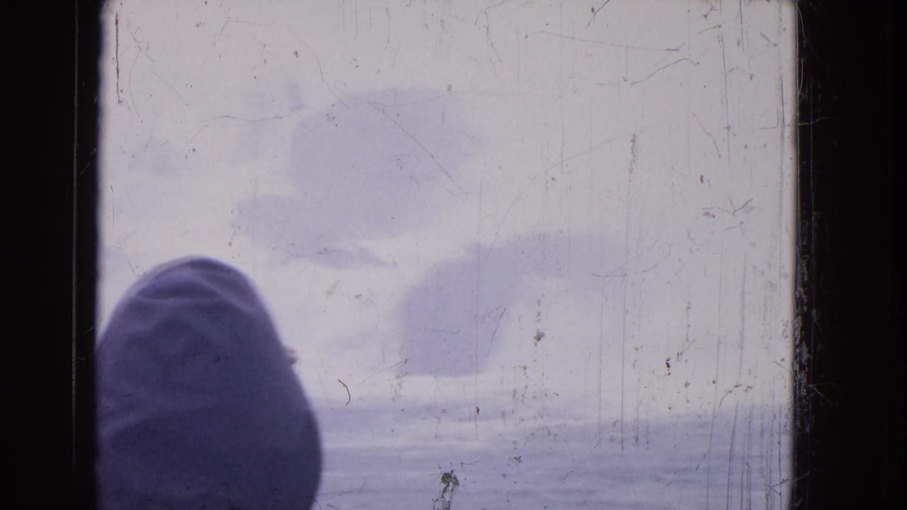 Hooded figure near a dark window frame with foggy water and pale sky outside.