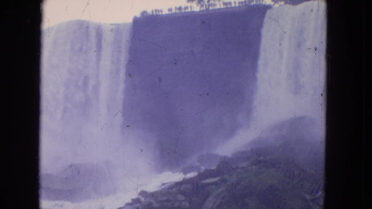 1964 Niagara Falls home-movie frame with a waterfall wall, foaming river, rocks, mist, and a distant overlook railing.
