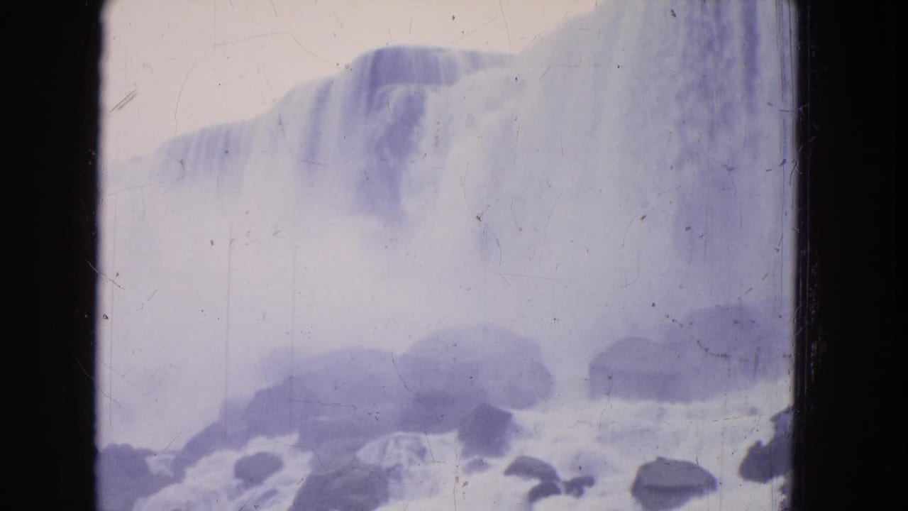 Mist-filled waterfall view with boulders and spray near the river edge.