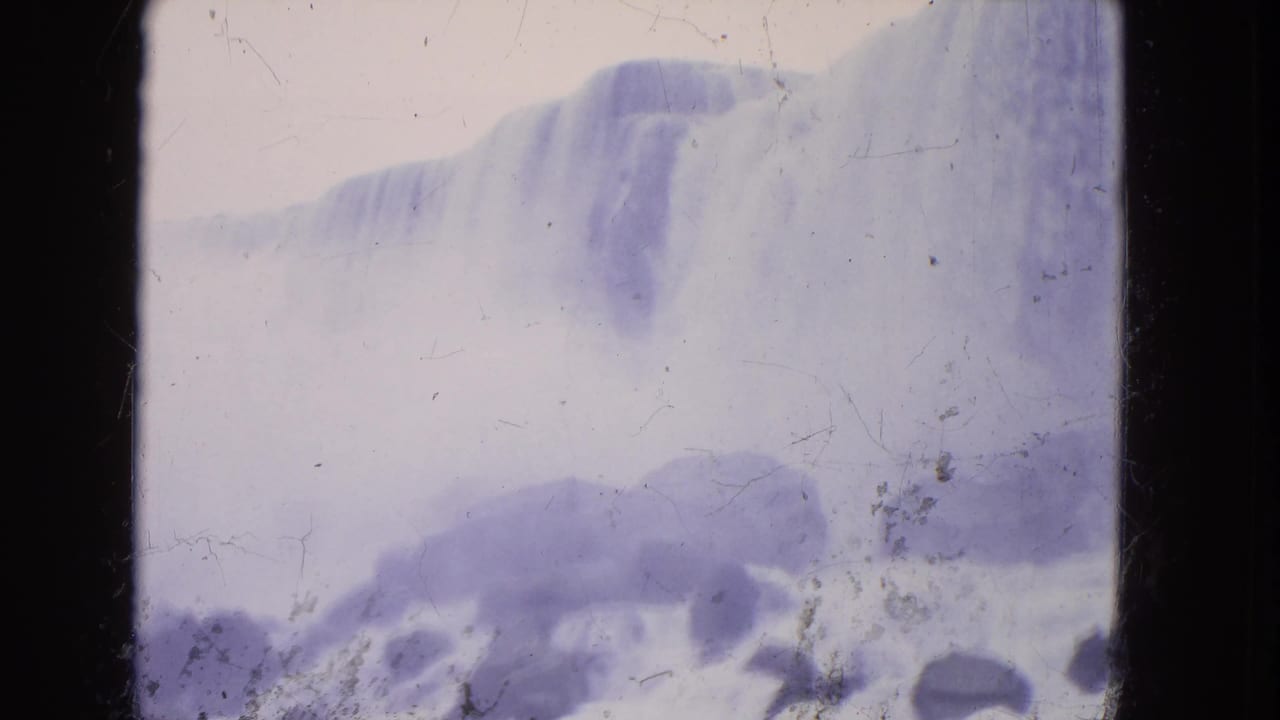 1964 8mm frame of Niagara Falls seen through haze, with a pale waterfall edge and dark rocks below.