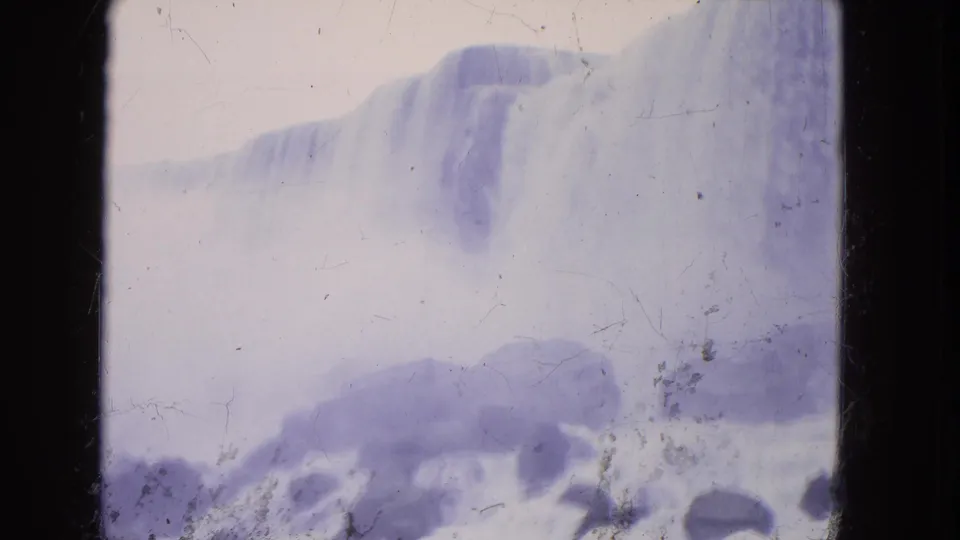 1964 8mm frame of Niagara Falls seen through haze, with a pale waterfall edge and dark rocks below.