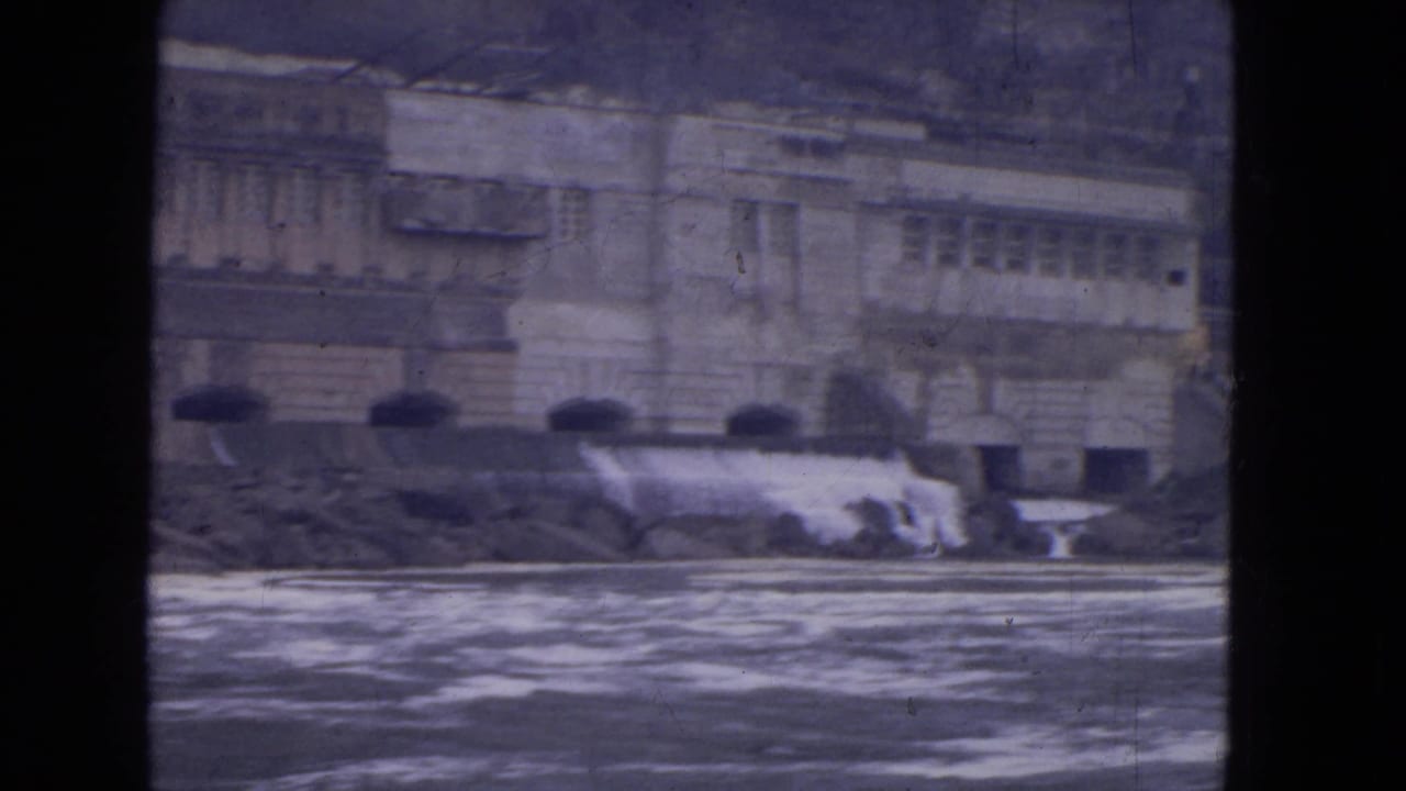 1964 Niagara Falls frame showing a large stone riverside building beyond churning water and a low cascade.