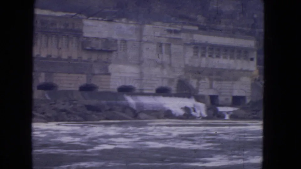 1964 Niagara Falls frame showing a large stone riverside building beyond churning water and a low cascade.
