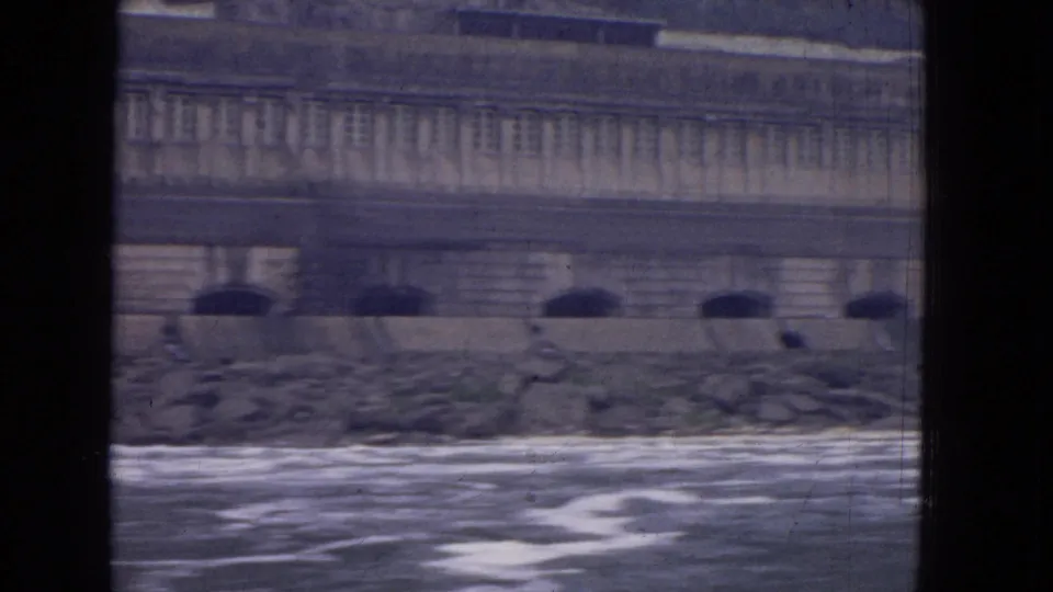 1964 Niagara River footage shows foamy water along a stone bank beneath a large weathered riverside structure.