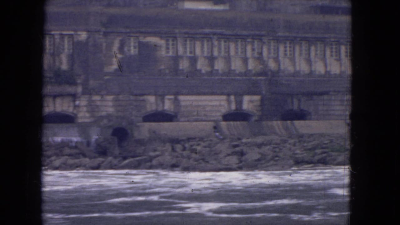 Soft 8mm frame from the Niagara River shows rough water, stone shoreline, and a long masonry building facade.
