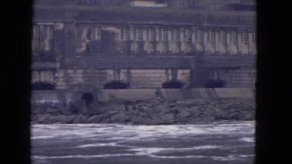 Soft 8mm frame from the Niagara River shows rough water, stone shoreline, and a long masonry building facade.