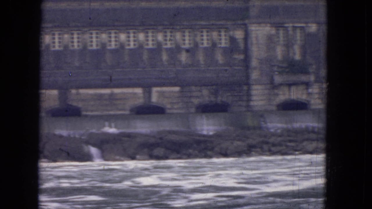 1964 8mm view near Niagara Falls showing a large riverside stone building beyond churning water, framed by dark window mullions.