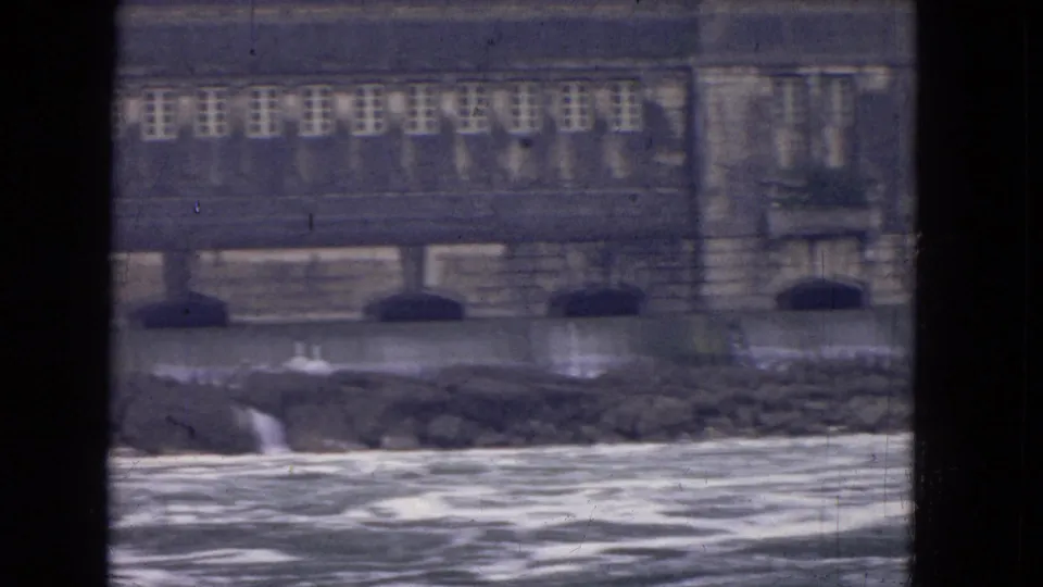1964 8mm view near Niagara Falls showing a large riverside stone building beyond churning water, framed by dark window mullions.