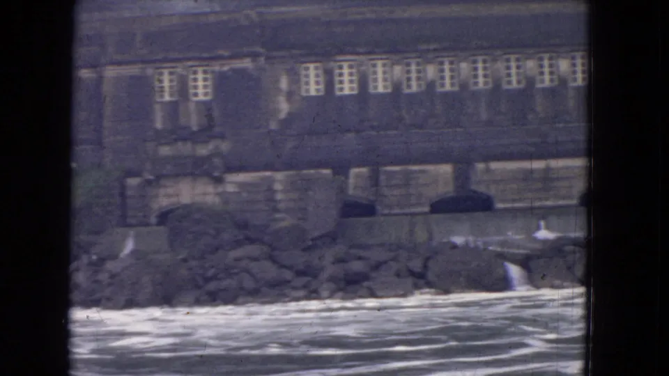 1964 8mm frame near Niagara Falls with a weathered stone building above rocks and moving river water, partly blocked by window framing.